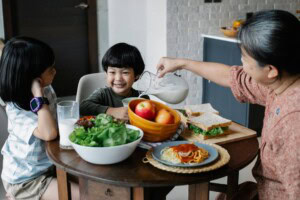 Grandmother pouring milk from jug in plate while sitting at served table with Asian boy and girl during breakfast in kitchen